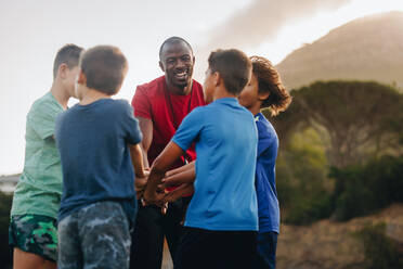 Rugbytrainer und seine Schüler fassen sich an den Händen. Sporttrainer, der seine Mannschaft auf einem Feld motiviert. Mann, der eine Gruppe von Grundschulkindern betreut. - JLPSF28911