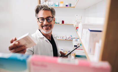 Pharmacist reading a medication label and holding a tablet in a chemist. Mature male healthcare worker filling online prescriptions in a pharmacy. - JLPSF28926