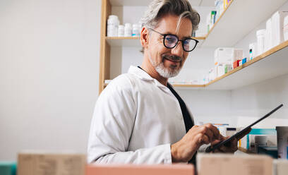 Mature pharmacist stocktaking with a tablet in a drug store. Senior man counting medical inventory using a checklist in a pharmacy. - JLPSF28927