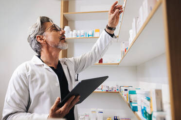 Senior pharmacist getting prescription medication from a shelf in a chemist. Male healthcare worker using a digital tablet to fill online prescriptions in a pharmacy. - JLPSF28929