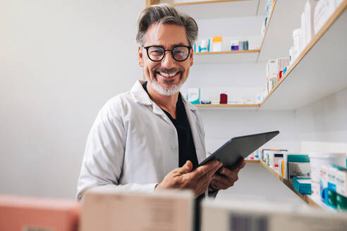 Glücklicher Apotheker, der eine Tablet in der Hand hält und in die Kamera lächelt, in einer Apotheke. Älteres medizinisches Personal, das Rezepte ausfüllt. - JLPSF28930