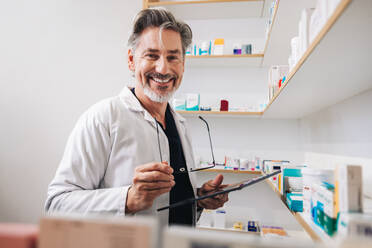 Senior pharmacist standing in a chemist and looking at the camera. Mature healthcare worker holding a digital tablet and eyeglasses in a pharmacy. - JLPSF28931