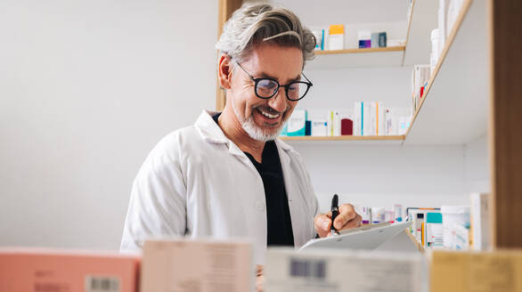 Ein männlicher Apotheker schreibt auf einem Klemmbrett, während er in einer Apotheke Rezepte ausstellt. Eine ältere medizinische Fachkraft arbeitet in einer Apotheke. - JLPSF28932