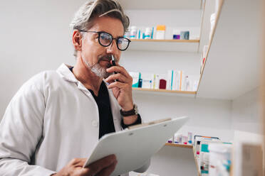 Senior man looking thoughtful while working in a pharmacy. Mature pharmacist stocktaking with a clipboard in a drug store. - JLPSF28933