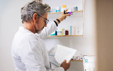 Male pharmacist processing a prescription in a drug store. Senior man reading a clipboard and getting medication from a dispensary shelf. - JLPSF28936