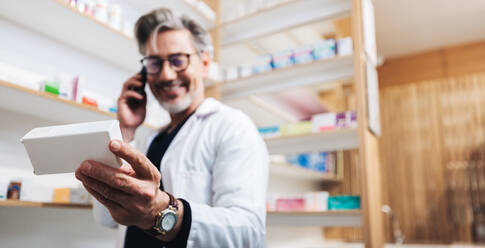 Apotheker in einer Drogerie, der eine Schachtel mit Tabletten in der Hand hält und mit einem Patienten telefoniert. Älteres medizinisches Personal liest das Etikett eines Medikaments, um ein Rezept zu bestätigen. Telefonanruf in einer Apotheke. - JLPSF28937
