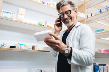 Pharmacist reading a medication label while talking to a patient on the phone. Senior healthcare worker confirming a prescription over a phone call. Mature man working in a clinic dispensary. - JLPSF28938