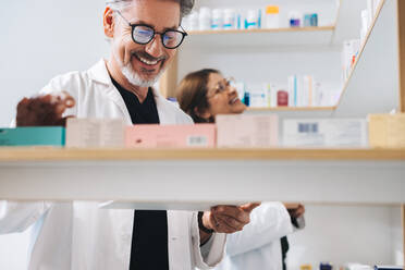 Male pharmacist picking prescription medication in a drug store. Happy senior man working in a pharmacy with his colleague. - JLPSF28956