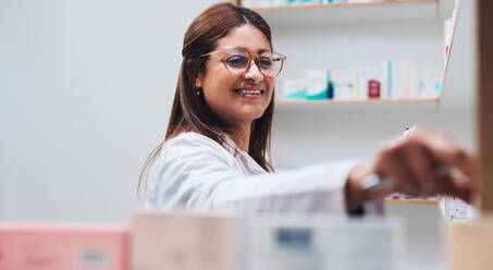 Pharmacy worker getting medication from a shelf. Indian female pharmacist dispensing a prescription in a chemist. Happy woman working in a drug store. - JLPSF28962