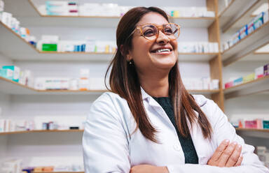 Happy female pharmacist standing in a drug store with crossed arms. Mature woman working in a pharmacy. Female healthcare worker in a chemist. - JLPSF28964