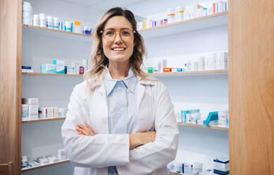 Portrait of a female pharmacist standing in a drug store. Female healthcare worker smiling at the camera with crossed arms. Woman working in a pharmacy. - JLPSF28969