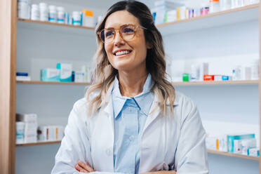 Female doctor standing in a pharmacy in a white coat. Female healthcare worker smiling with crossed arms. Woman working in a hospital dispensary. - JLPSF28970