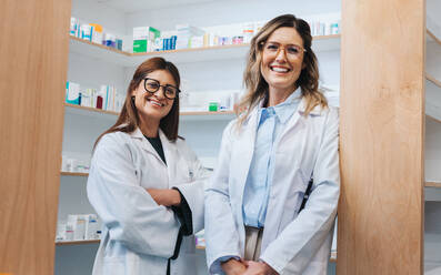 Female pharmacists standing in a chemist and smiling at the camera. Two healthcare professionals working together in a pharmacy. - JLPSF28972