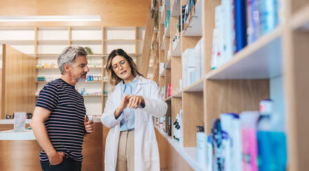 Female pharmacist assisting a man in a drug store. Healthcare professional showing a skincare product to a patient in a pharmacy. - JLPSF28975