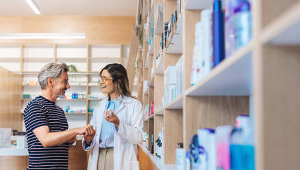 Friendly pharmacist assisting a man in a chemist. Female healthcare professional showing a skincare product to a patient in a pharmacy. - JLPSF28976