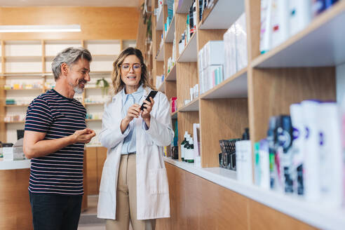 Apothekerin zeigt einem Mann ein Hautpflegeprodukt in einer Apotheke. Mitarbeiterin des Gesundheitswesens hilft einem Patienten in einer Apotheke. - JLPSF28977