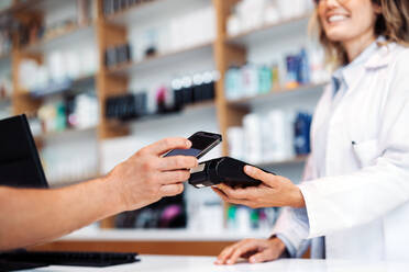 Apothekerin beim Scannen eines Mobiltelefons an einem Kartenautomaten in einer Drogerie. Mitarbeiter des Gesundheitswesens beim Empfang einer nfc-Zahlung für Medikamente in einer Apotheke. - JLPSF28981