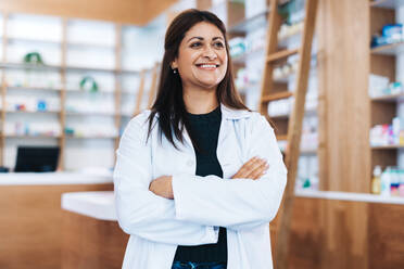 Indian female pharmacist standing in a chemist with crossed arms. Female healthcare professional smiling and looking away. Woman working in a drug store. - JLPSF28985