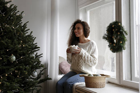 Glückliche Frau auf der Fensterbank sitzend mit einer Tasse Marshmallow-Kakao zu Hause - SYEF00156