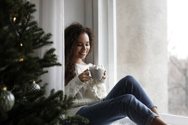 Happy young woman sitting on window sill with cup of marshmallow cocoa at home - SYEF00157