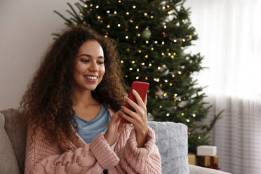 Glückliche junge Frau mit Smartphone vor dem Weihnachtsbaum zu Hause - SYEF00162