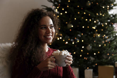 Lächelnde junge Frau mit einer Tasse Marshmallow-Kakao vor einem Weihnachtsbaum sitzend - SYEF00166