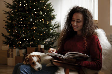 Happy woman sitting with dog and reading book at home - SYEF00170