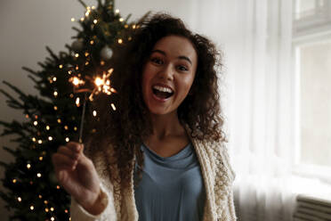 Fröhliche junge Frau mit Wunderkerze vor einem Weihnachtsbaum stehend - SYEF00175