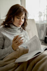 Junge Frau mit Kaffeetasse, die zu Hause ein Buch liest - ANAF00750