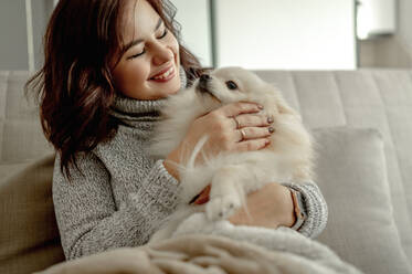 Cheerful young woman stroking Pomeranian dog at home - ANAF00752