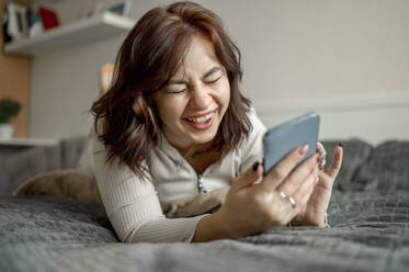 Fröhliche junge Frau mit Smartphone, die zu Hause auf dem Bett liegt - ANAF00754