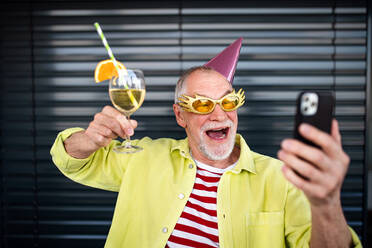 Portrait of crazy senior man with party hat and drink standing outdoors, taking selfie. - HPIF05361