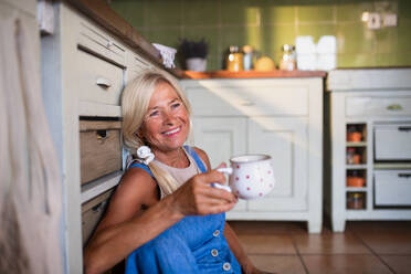Eine glückliche ältere Frau sitzt auf dem Boden und trinkt drinnen in der Küche Tee. - HPIF05380