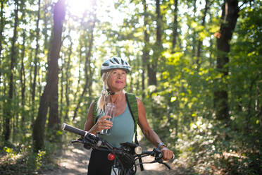 Eine glückliche ältere Frau Radfahrer trinken Wasser aus der Flasche im Freien im Wald. - HPIF05381