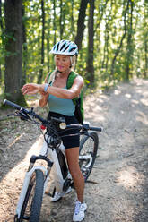 An active senior woman biker sitting on bicycle and setting smartwatch outdoors in forest. - HPIF05384
