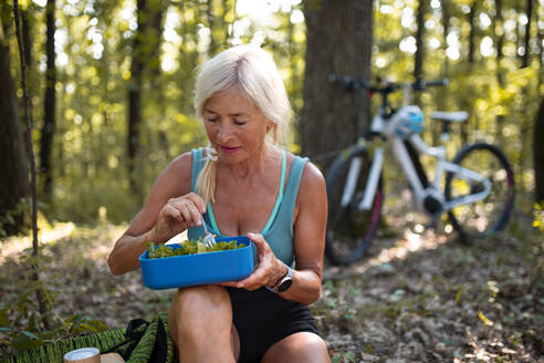 Eine glückliche ältere Frau Biker ruht, sitzen und essen Snack im Freien im Wald. - HPIF05387