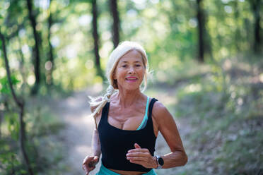 A happy active senior woman jogging outdoors in forest, waist up. - HPIF05399