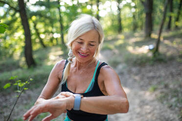 Porträt einer aktiven älteren Läuferin, die im Freien im Wald steht und ihre Smartwatch einstellt. - HPIF05401