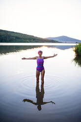 A portrait of active senior woman swimmer standing and stretching outdoors in lake. - HPIF05418