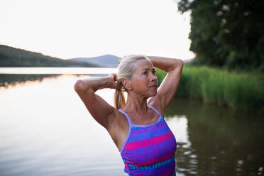 A portrait of active senior woman swimmer standing and stretching outdoors in lake. - HPIF05419