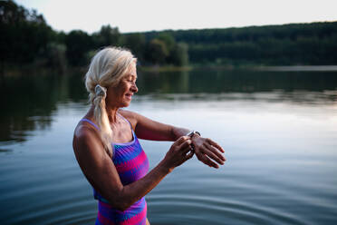 Ein Porträt einer aktiven älteren Schwimmerin, die ihre Smartwatch im Freien in einem See einstellt. - HPIF05423