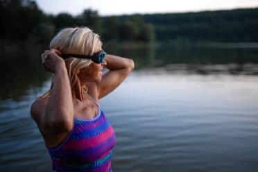 Ein Porträt einer aktiven älteren Schwimmerin, die im Freien in einem See steht und ihre Schwimmbrille anlegt. - HPIF05434
