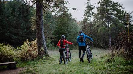 Rückansicht eines älteren Radfahrerehepaars, das zu Fuß unterwegs ist und E-Bikes im Herbst im Wald schiebt. - HPIF05456