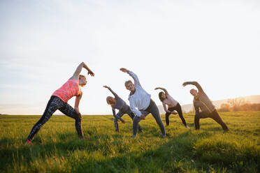 Eine Gruppe von Senioren mit einem Sportlehrer, die bei Sonnenuntergang draußen in der Natur Sport treiben, aktiver Lebensstil. - HPIF05513