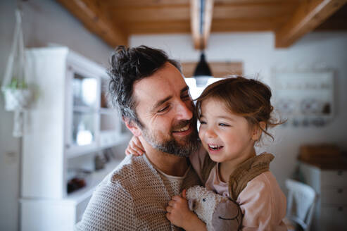 A mature father with small daughter standing indoors at home, holding and hugging. - HPIF05524