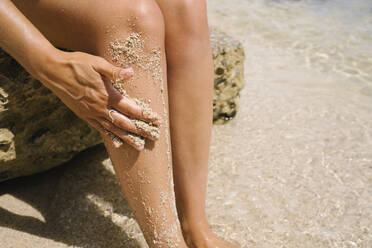 Frau, die ihre Beine mit Sand peelt, sitzt auf einem Felsen am Strand - MMPF00575