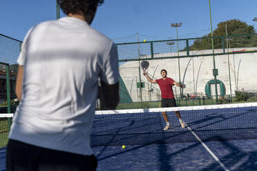 Junger Mann mit Freund spielt Paddle-Tennis auf einem Sportplatz an einem sonnigen Tag - FMOF01606