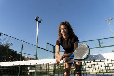 Junge Frau mit Tennisschläger, die sich auf dem Tennisplatz an das Netz lehnt - FMOF01619