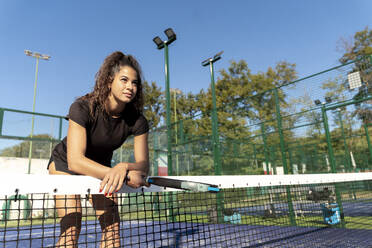 Junge Frau mit Tennisschläger am Tennisnetz lehnend - FMOF01620