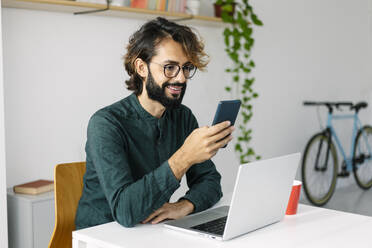 Happy bearded businessman using smart phone at desk in office - XLGF03126